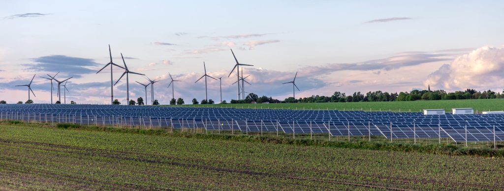 A field near solar panels, and wind turbines in the horizon